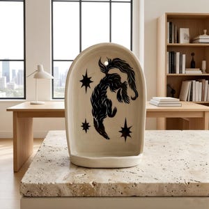 May include: Cream ceramic wall hanging featuring a black illustration of a stylized animal and stars. The arched piece has a small shelf at the bottom and a hole at the top for hanging. Decorative home decor.