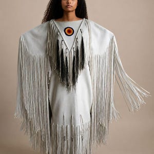 May include: A light gray dress with long fringe sleeves and hem. The dress features a beaded circular design at the neckline and black fringe accents. The model is wearing the dress with arms outstretched.