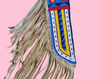 May include: A beaded, fringed item with a tan suede fringe and a colorful beaded section. The beaded section features blue, red, yellow, and white beads in geometric patterns. The fringe hangs from the side of the beaded section.