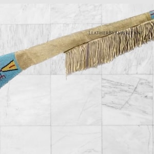 Peut inclure: Long objet en cuir beige avec des franges et des détails perlés. Les extrémités présentent des sections perlées bleu clair avec des motifs géométriques jaunes, rouges et noirs. Le texte "LEATHERBYAMUKELAN" est visible.