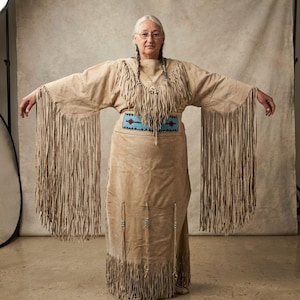 May include: A woman wearing a tan suede dress with long fringe sleeves and a fringed hem. The dress features a turquoise and black beaded belt and turquoise beads hanging from the skirt. The woman is wearing glasses and has gray hair.