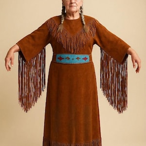 Puede incluir: Vestido de ante marrón con flecos en el escote, las mangas y el dobladillo. El vestido presenta un cinturón de cuentas turquesa y rojo. Las mangas son largas y acampanadas, y el vestido tiene un escote redondo.