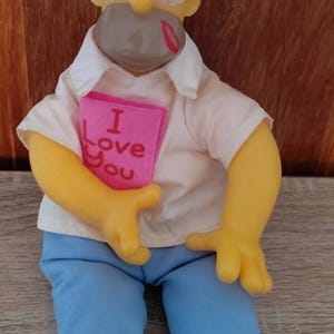 May include: A yellow cartoon character doll with large white eyes, wearing a white collared shirt and light blue trousers. The doll has red lipstick kiss marks on its forehead and chin, and holds a pink rectangular sign that says I Love You.