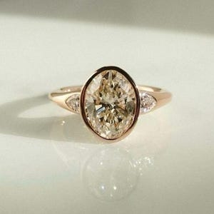 May include: A rose gold ring with a large, oval-cut diamond in a champagne colour. The diamond is bezel-set and has smaller, triangular accent stones. The ring is shown on a reflective surface.