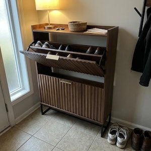 May include: A dark brown wooden shoe cabinet with a flip-down door and a top shelf. The cabinet has a textured front and a black metal base. Several pairs of shoes are visible inside the cabinet. A lamp and a basket are on the top shelf.