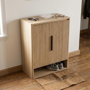 May include: A light-colored wooden shoe cabinet with a vertical grooved design on the door. The cabinet has a black handle and a lower open shelf with two pairs of shoes. Keys, glasses, and papers rest on top. A woven mat sits on the floor.
