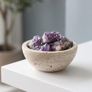 Peut inclure: Un petit bol rond en travertin beige à la surface poreuse et texturée, rempli de plusieurs amas de cristaux d'améthyste violet foncé aux bords irréguliers. Le bol présente un bord lisse et incurvé et repose sur une surface blanche plane.