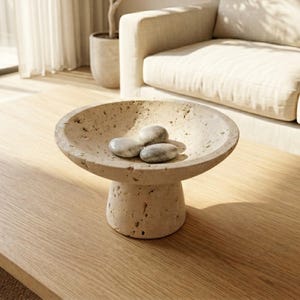 May include: A beige travertine pedestal bowl with a porous, textured surface and a wide, shallow basin. Three smooth, rounded grey and white river stones are placed inside the center of the bowl, resting on the bottom of the interior surface.