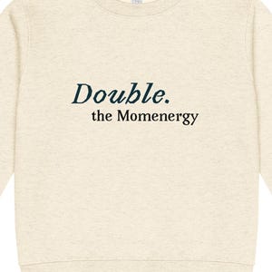 Könnte beinhalten: Ein hellbeige Sweatshirt mit dem Text "Double. the Momenenergy" in Dunkeltürkis. Das langärmlige Oberteil mit Rundhalsausschnitt ist aus einem weichen Material gefertigt.