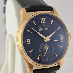 May include: A gold-tone wristwatch with a dark blue dial, black leather strap, and gold hands. The dial features the text Jaeger-LeCoultre, 12, 9, 3, 2029, JUL, and a moon phase sub-dial with a sun icon. The outer edge displays numbers 1 through 31.