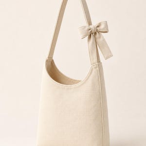 May include: A cream-colored canvas hobo bag featuring a single shoulder strap and a decorative fabric bow attached to the upper side of the strap. The bag has a curved top opening and a structured, vertical rectangular body design.