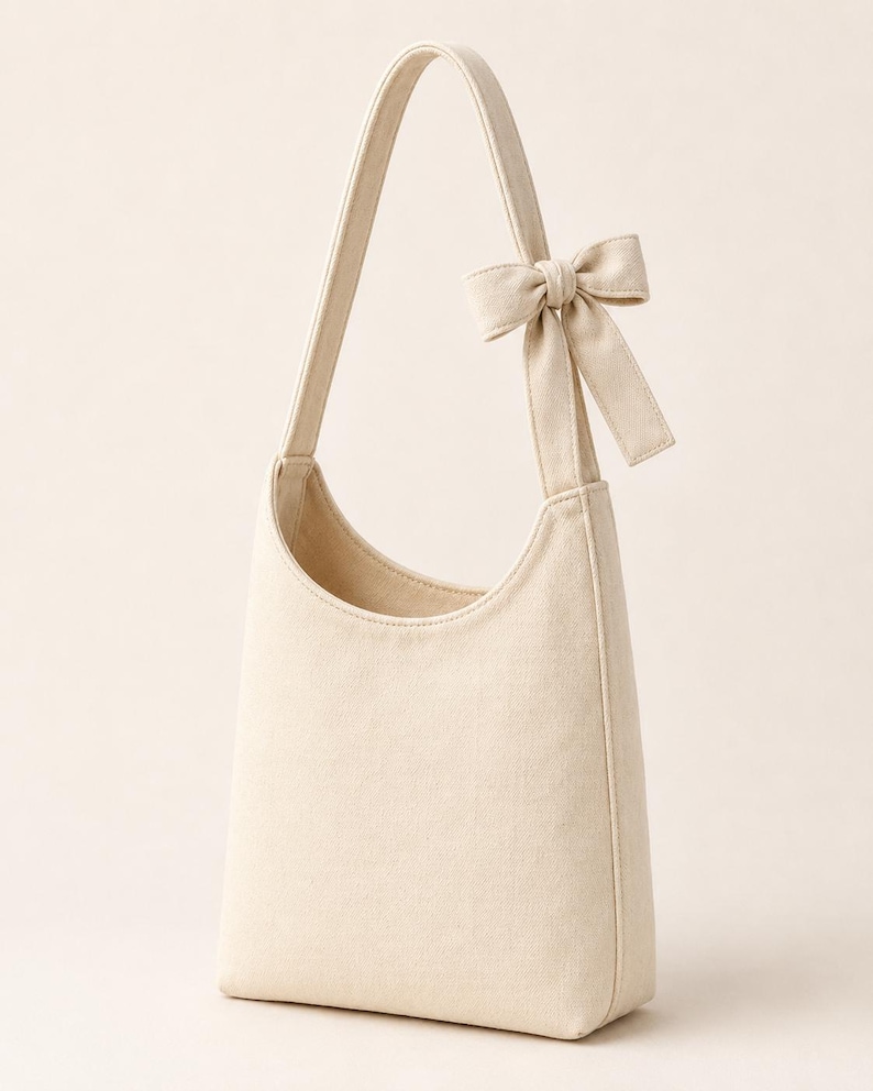 May include: A cream-colored canvas hobo bag featuring a single shoulder strap and a decorative fabric bow attached to the upper side of the strap. The bag has a curved top opening and a structured, vertical rectangular body design.