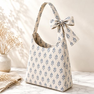 May include: A cream-colored handbag featuring a repeating blue botanical print of small sprigs. The bag has a single shoulder strap and is accented with a matching fabric bow tied to the top of the strap, creating a cohesive and structured appearance.