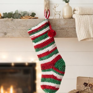 Puede incluir: Un calcetín navideño tejido a mano con rayas horizontales en colores alternos rojo, blanco y verde. El calcetín incluye un puño grueso doblado en la parte superior, un pequeño pompón rojo unido al lateral y un lazo para colgar a juego.