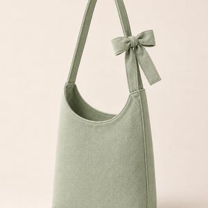 May include: A sage green hobo-style shoulder bag crafted from a textured fabric. The bag features a single integrated shoulder strap adorned with a decorative fabric bow detail on one side, a curved top opening, and a structured, vertical silhouette.