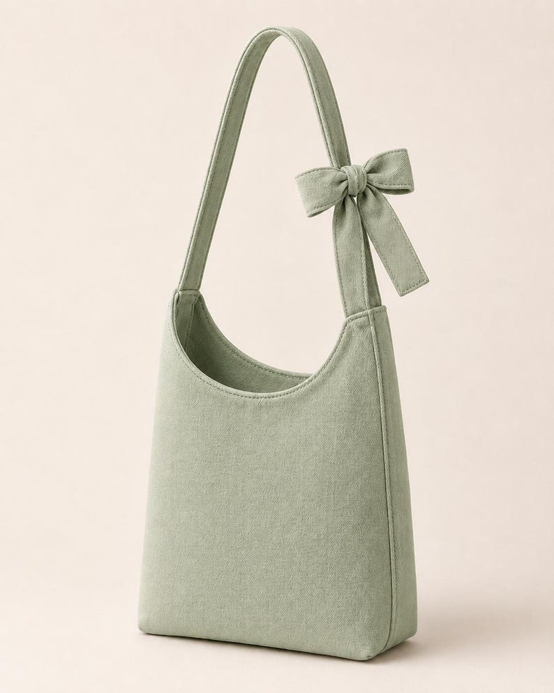 May include: A sage green hobo-style shoulder bag crafted from a textured fabric. The bag features a single integrated shoulder strap adorned with a decorative fabric bow detail on one side, a curved top opening, and a structured, vertical silhouette.