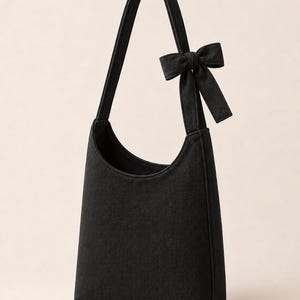May include: A black hobo-style shoulder bag featuring a single strap and a decorative fabric bow attached to the upper side. The bag has a curved top opening and a structured, vertical rectangular body made of a solid black textile material.