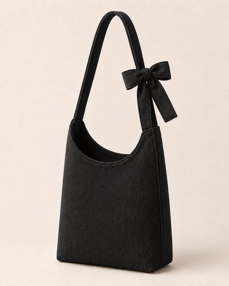 May include: A black hobo-style shoulder bag featuring a single strap and a decorative fabric bow attached to the upper side. The bag has a curved top opening and a structured, vertical rectangular body made of a solid black textile material.