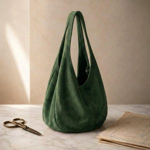 May include: A deep forest green hobo bag crafted from a soft, matte suede material. The bag features a slouchy, unstructured silhouette with a single integrated shoulder strap and a wide, open top opening that tapers toward the base.