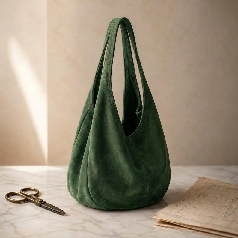 May include: A deep forest green hobo bag crafted from a soft, matte suede material. The bag features a slouchy, unstructured silhouette with a single integrated shoulder strap and a wide, open top opening that tapers toward the base.