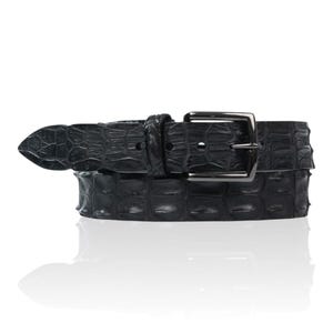 Op de afbeelding: Een zwarte leren riem met een textuur die op krokodillenleer lijkt. De riem heeft een zilverkleurige rechthoekige gesp en een puntig uiteinde. De riem wordt getoond tegen een witte achtergrond.