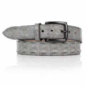 Op de afbeelding: Een grijze riem met een textuur die op krokodillenleer lijkt. De riem heeft een zwarte metalen gesp en een puntige punt. De riem wordt gepresenteerd op een witte achtergrond.