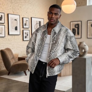 Peut inclure: Une veste à manches longues avec un motif peau de serpent gris et blanc, portée sur un débardeur blanc uni et un jean noir. La veste présente un col classique, une fermeture éclair sur le devant et des manches longues avec poignets.