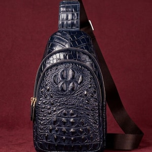 May include: A dark blue sling bag featuring a textured crocodile skin pattern. The bag has a rounded top, a front zippered pocket, and a dark brown adjustable fabric strap with a silver-toned metal buckle attached to the top of the bag.