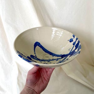 Könnte beinhalten: Eine Keramikschale mit glänzend cremefarbener Oberfläche und einem handgemalten abstrakten Design in Kobaltblau. Das Muster besteht aus kräftigen Pinselstrichen und Farbspritzern, die sich vom Boden des Inneren bis zum Rand erstrecken.