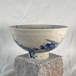 Könnte beinhalten: Eine Keramikschale mit cremefarbener Basis und einem blauen Sprenkelmuster. Die Schale hat einen breiten, ausgestellten Rand, der sich zu einem schmalen, erhöhten runden Fuß verjüngt, und ist mit einer glatten, glasierten Oberfläche versehen.