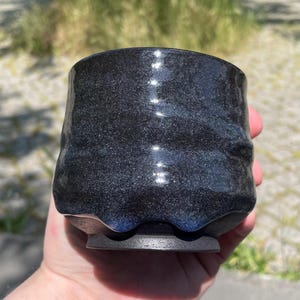 Könnte beinhalten: Eine handgefertigte, dunkelblaue und schwarze Keramiktasse mit strukturierter Oberfläche. Die Tasse hat eine einzigartige, unregelmäßige Form und einen kleinen, dunklen Boden. Der Innenraum ist tiefblau, die Außenseite hat ein gesprenkeltes, fast galaxieartiges Aussehen.