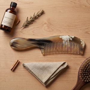 Puede incluir: Un peine de cuerno con mango curvo y dientes finos, exhibido con una botella de aceite para el cabello, un paño de lino doblado, un cepillo de pelo de madera, romero y palitos de canela. La botella tiene una etiqueta que dice "Serene Horn Premium Hair Oil (Argan & Sandalwood)".