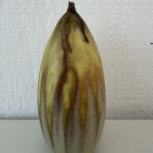 Op de afbeelding: Een hoge, traanvormige keramische vaas met een gladde, lichtgele basisglazuur en verticale, druipende strepen in donkerbruine en aardse tinten. De vaas loopt taps toe naar een smalle, puntige hals en heeft een rond, bolvormig lichaam.