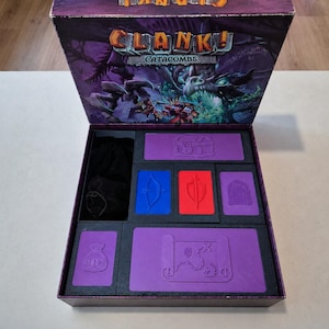 May include: The Clank! Catacombs board game box is open, revealing a black plastic insert with purple, blue, and red storage compartments. The purple lid features embossed icons of a treasure chest, a map, and a coin bag. A black cloth bag is included.