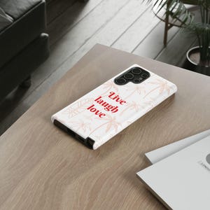 May include: A white smartphone case featuring a repeating pattern of light orange palm trees and a surfboard. The text Live laugh love is printed in red on the back of the case, which also includes a black camera cutout with multiple lenses.