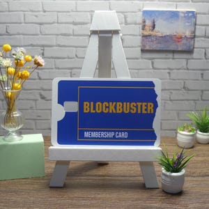 A blue and yellow Blockbuster Membership Card decal is displayed upright on a white easel, with a vase of flowers and small potted plants in the background.
