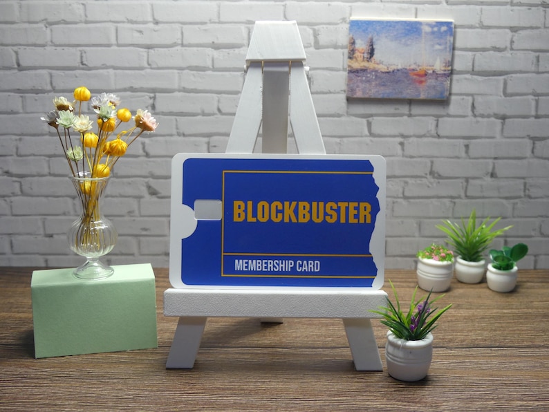 A blue and yellow Blockbuster Membership Card decal is displayed upright on a white easel, with a vase of flowers and small potted plants in the background.