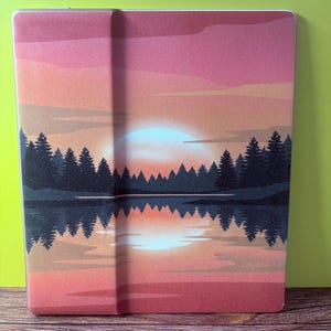 May include: A rectangular e-reader cover featuring a printed sunset landscape. The design displays a bright sun setting behind a dark silhouette of pine trees, with the reflection of the trees and the sky visible in the water below in shades of pink.