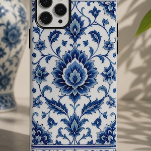 以下が含まれることがあります： 白い背景に青い花柄のダマスク模様が繰り返されるスマートフォンケース。複雑で対称的な植物のモチーフと葉がデザインされており、長方形のケースの左上隅には黒いカメラ用の切り抜きがあります。