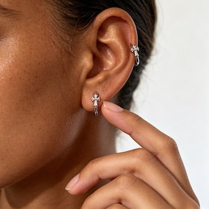 May include: Two silver-toned hoop earrings, each featuring a small, detailed cross charm attached to the front. One earring is positioned in the earlobe, while the other is placed higher up on the cartilage, both displaying a polished metallic finish.