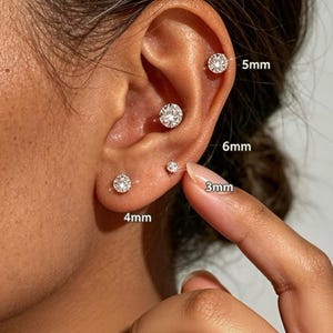 Könnte beinhalten: Vier runde, klare, facettierte Ohrstecker in silberfarbenem Metall, die an einem Ohr gezeigt werden. Die Ohrringe sind mit ihren jeweiligen Größen beschriftet: 3mm, 4mm, 5mm und 6mm, was den Durchmesser der einzelnen Steinfassungen angibt.