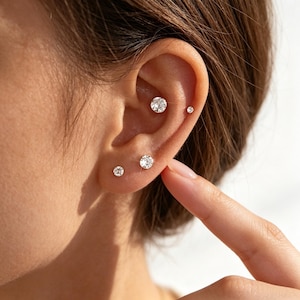 May include: An ear with four small, clear crystal stud earrings of varying sizes. The largest earring is at the top, with the smallest at the bottom. The earrings are set against a soft, neutral background. The image is a close-up.