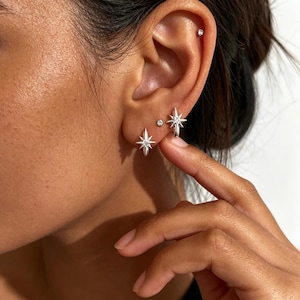 May include: A set of silver-toned earrings featuring a starburst design with a central clear stone. The jewelry includes two star-shaped studs and a small circular stud, all worn on the earlobe, plus a single small circular stud in the upper cartilage.