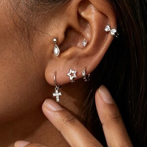 Pode incluir: Uma orelha é adornada com vários brincos de prata. Os brincos incluem um brinco em forma de gota, um pequeno brinco de diamante, um brinco em forma de estrela, um pequeno aro, um pingente de cruz e uma argola de orelha em forma de laço. Todos os brincos são prateados e com pequenas pedras preciosas transparentes.
