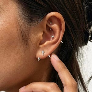 May include: A silver-toned ear piercing set featuring a small cross stud earring in the lobe, flanked by two conical spike studs. The upper ear cartilage displays two additional silver-toned conical spike piercings arranged vertically.