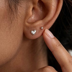 Puede incluir: Primer plano de una oreja con dos pendientes de plata. Uno tiene forma de corazón pequeño, el otro es una pequeña bola de plata. Un dedo toca suavemente la oreja. Los pendientes son probablemente de plata de ley.