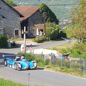 Peut inclure: Une voiture de course bleue, avec un pilote portant un casque, roule sur une route goudronnée. Le véhicule à toit ouvert est équipé d'un aileron arrière proéminent et de détails blancs. La scène se déroule dans une zone rurale avec des bâtiments et des collines verdoyantes.