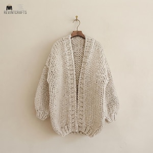 Peut inclure: Un cardigan en tricot épais blanc cassé avec des manches longues et une ouverture sur le devant. Le vêtement présente un motif de maille texturé sur toute la surface, incluant une bordure côtelée le long de l'ouverture et de l'ourlet.