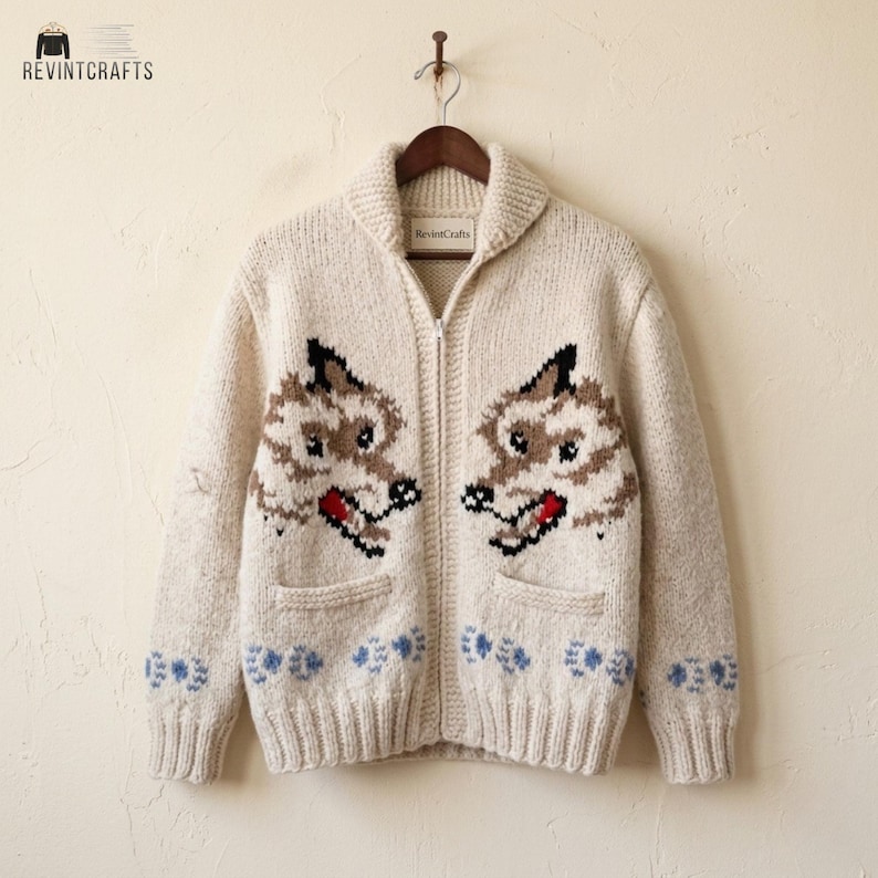 May include: A cream-colored knit zip-up cardigan featuring a shawl collar and two front pockets. The sweater displays two brown and black wolf head motifs on the chest and a repeating pattern of small blue paw prints along the bottom hem.