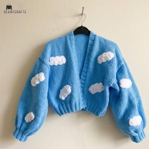 Può includere: Un cardigan in maglia azzurro con scollo a V, maniche lunghe con polsini a coste e orlo corto. Il capo è decorato con diverse applicazioni a forma di nuvola bianche, testurizzate e tridimensionali sul davanti e sulle maniche.